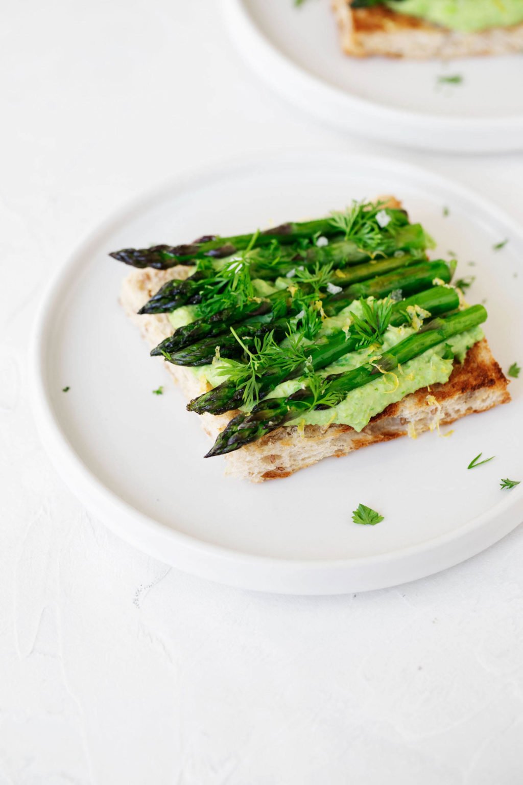 An angled photograph of a tartine made with green pea hummus and grilled asparagus spears. It rests on a white plate and white surface.