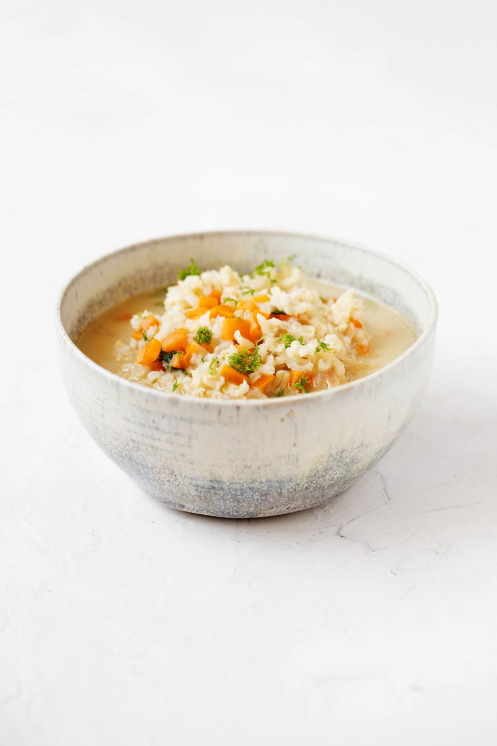 An angled photo of a ceramic bowl, filled with rice, broth, and vegetables. It rests on a white surface.