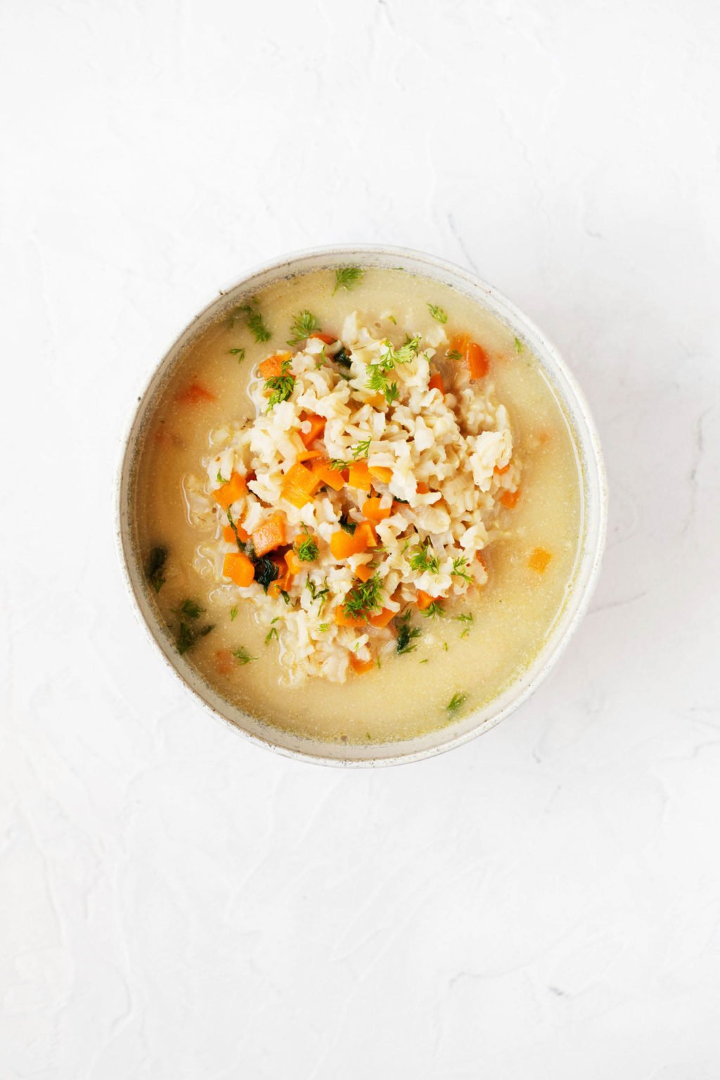 An overhead image of a white bowl on a white surface. It contains a portion of vegan avgolemono soup with rice and dill.