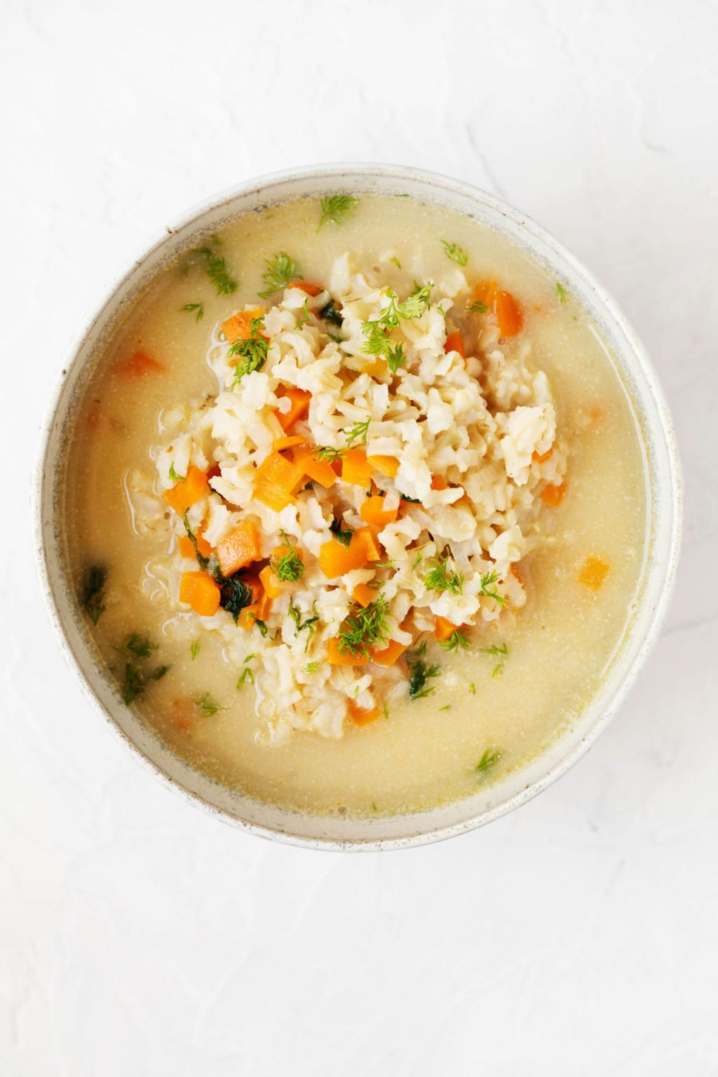 An overhead image of a white bowl, containing vegan avgolemono soup and flecked with green dill.