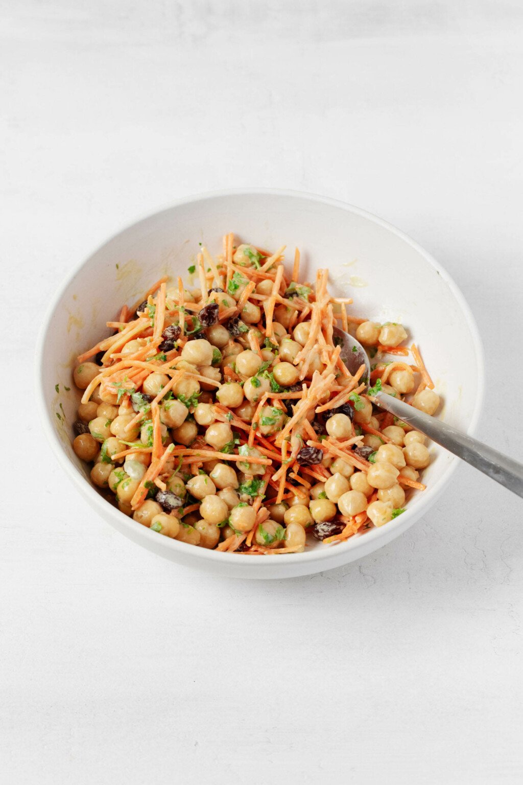 An overhead image of a creamy mixture of chickpeas, raisins, parsley, and grated carrots in a white mixing bowl. 