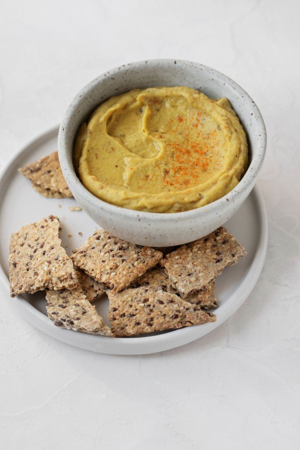 A small white dip bowl contains a light orange dip. It's paired with seedy crackers.