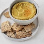 A small white dip bowl contains a light orange dip. It's paired with seedy crackers.