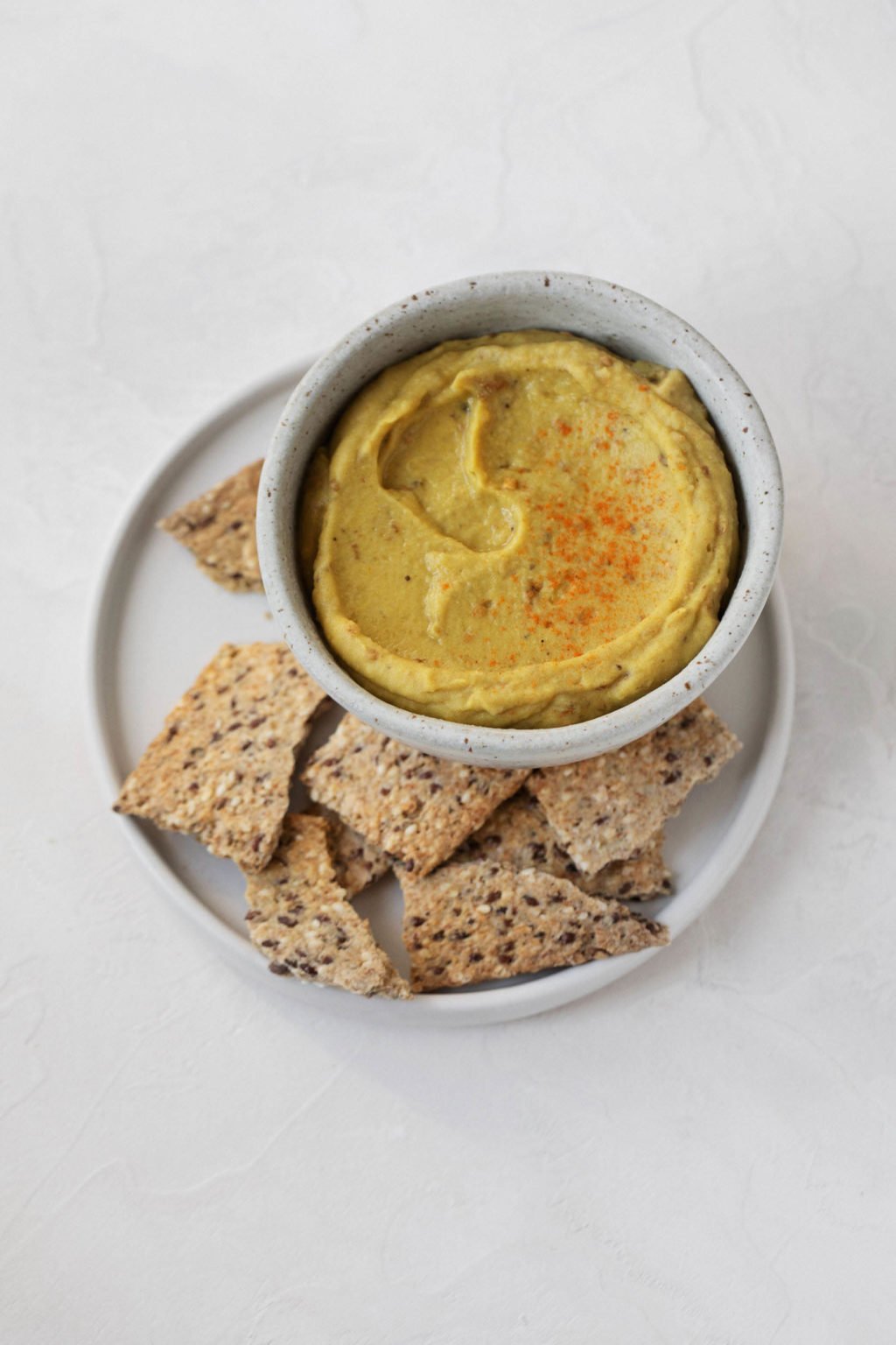 An overhead image of a white surface, with a white bowl filled with smoky eggplant red lentil dip and crackers on top.