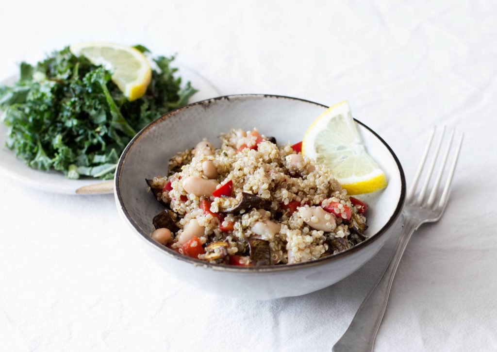 A bowl of cooked quinoa, eggplant, beans, and peppers is accompanied by a small plate of greens. They rest on a white tablecloth.