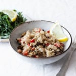 A bowl of cooked quinoa, eggplant, beans, and peppers is accompanied by a small plate of greens. They rest on a white tablecloth.