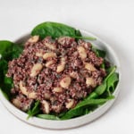 A white, rimmed plate is covered in green spinach leaves and a red quinoa bean salad. It rests on a white surface.