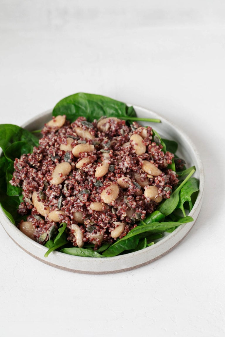 A white, rimmed plate is covered in green spinach leaves and a red quinoa bean salad. It rests on a white surface.