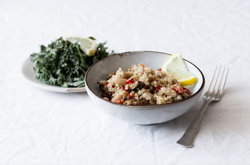 An eggplant quinoa dish with red bell pepper, lemon, and beans has been piled into a small white bowl with a brown rim. There's a plate of steamed greens nearby.