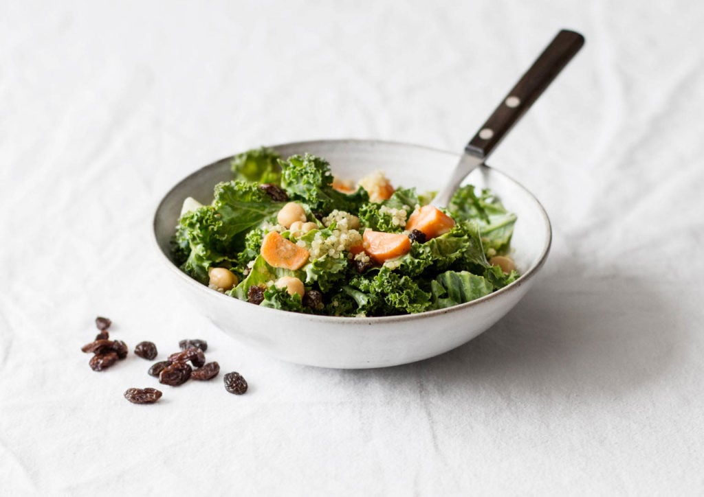 A white ceramic bowl rests on a white tablecloth. It contains a kale salad with a creamy dressing.