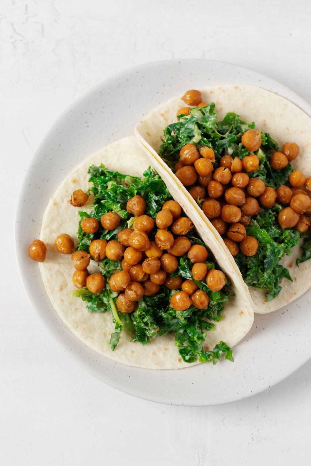 Two creamy kale and crispy chickpea tacos have been arranged on a round, white rimmed plate. 