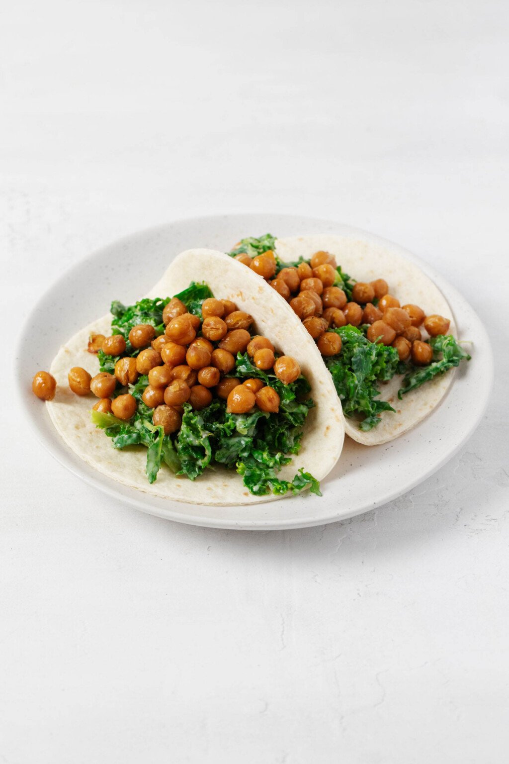 Two creamy kale and crispy chickpea tacos have been arranged on a round, white rimmed plate.