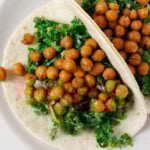 Two creamy kale and crispy chickpea tacos have been arranged on a round, white rimmed plate.