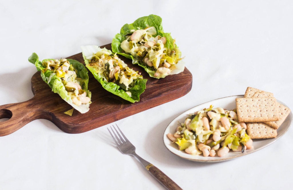A small, wood cutting board has been topped with plant-based lettuce wraps. A dish of warm legumes and crackers rests nearby.