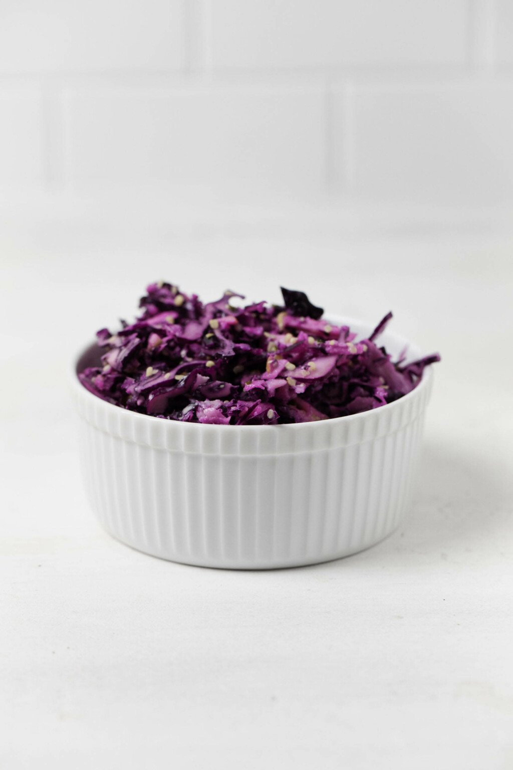 An apple cabbage slaw with tahini has been placed in a small, round white bowl. It rests against a white backdrop.