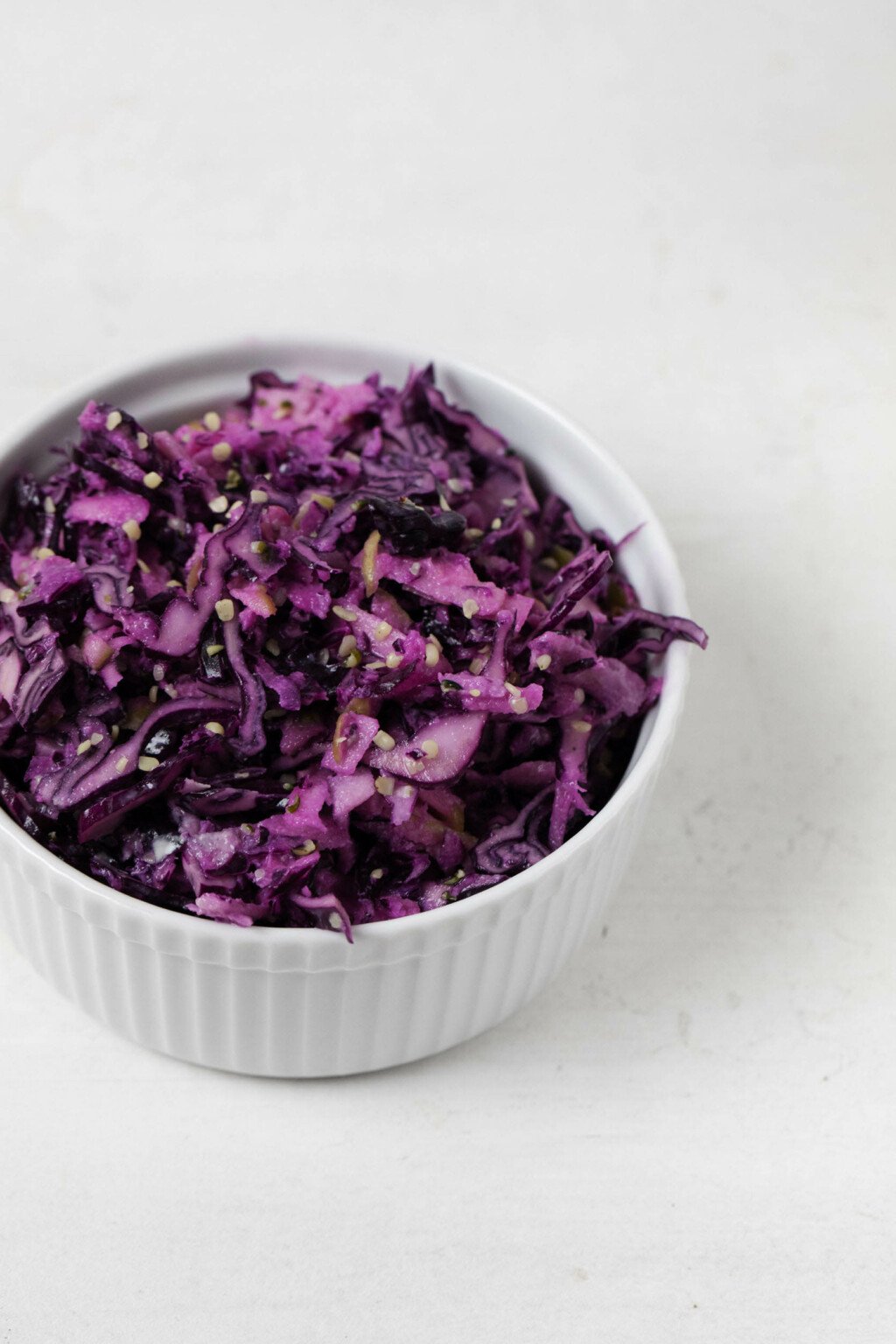 A white, rimmed bowl holds a mix of red cabbage, hemp seeds, and apple. It rests on a white surface.