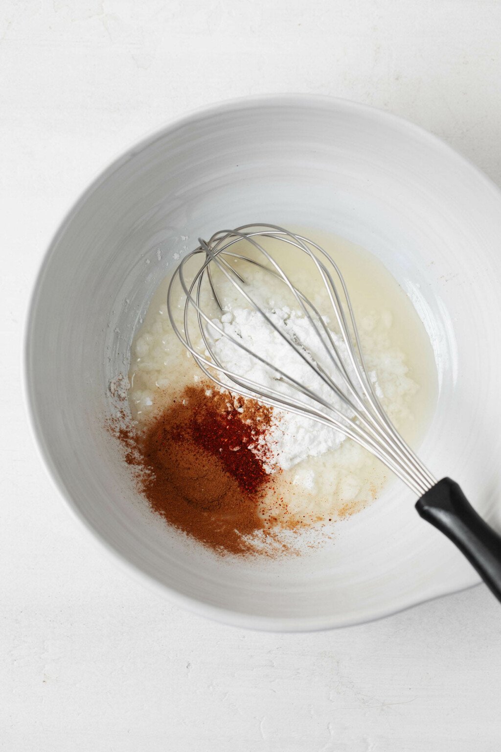 A white mixing bowl is filled with beaten aquafaba and spices. A whisk is resting in the bowl.