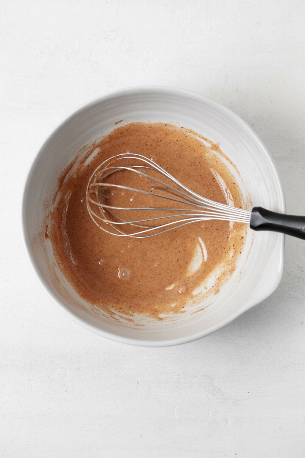 A white mixing bowl rests on a white surface with a whisk resting along its side. It's filled with a sticky, brown liquid mixture.