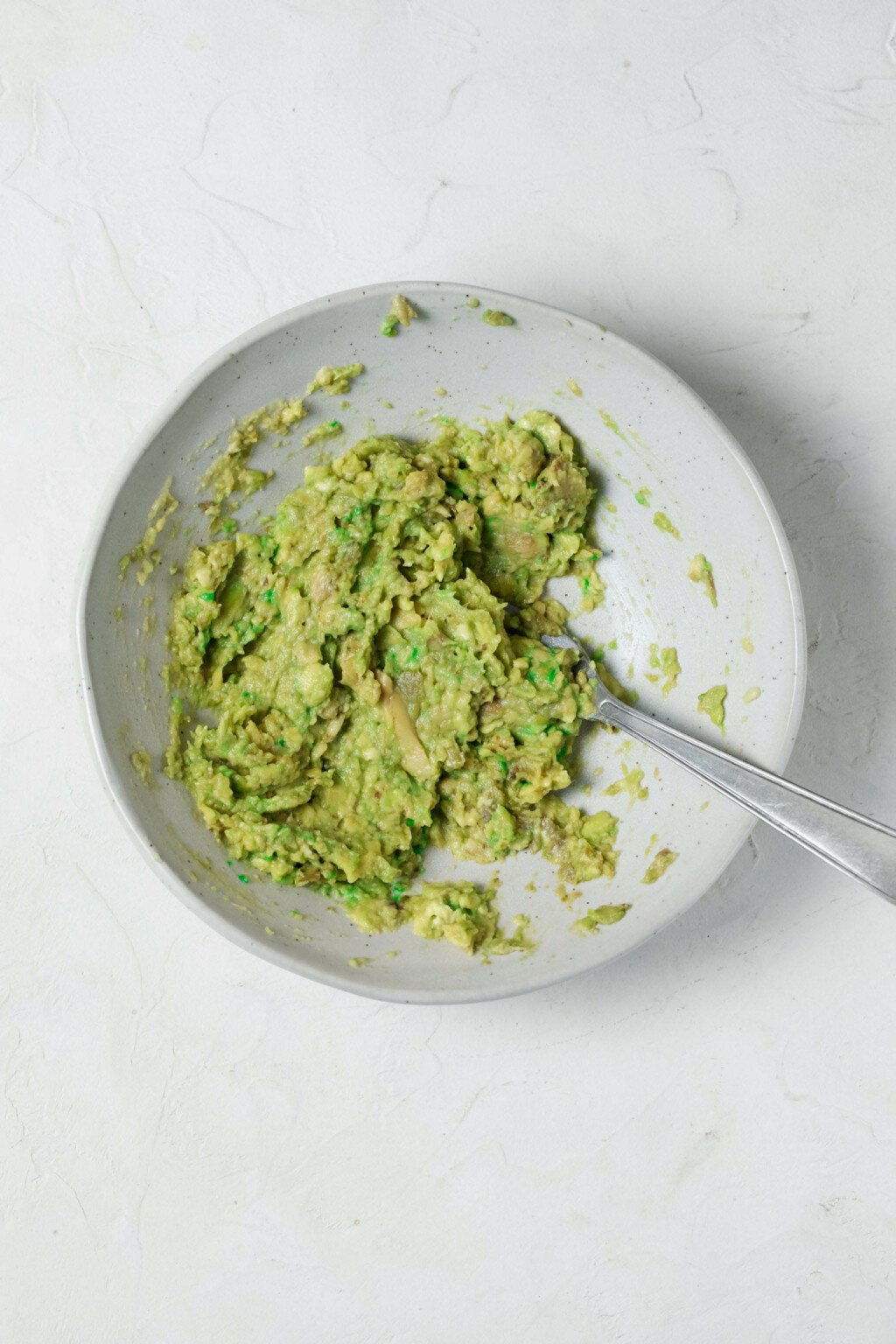 An overhead image of a white bowl, which contains pale green, mashed avocado.