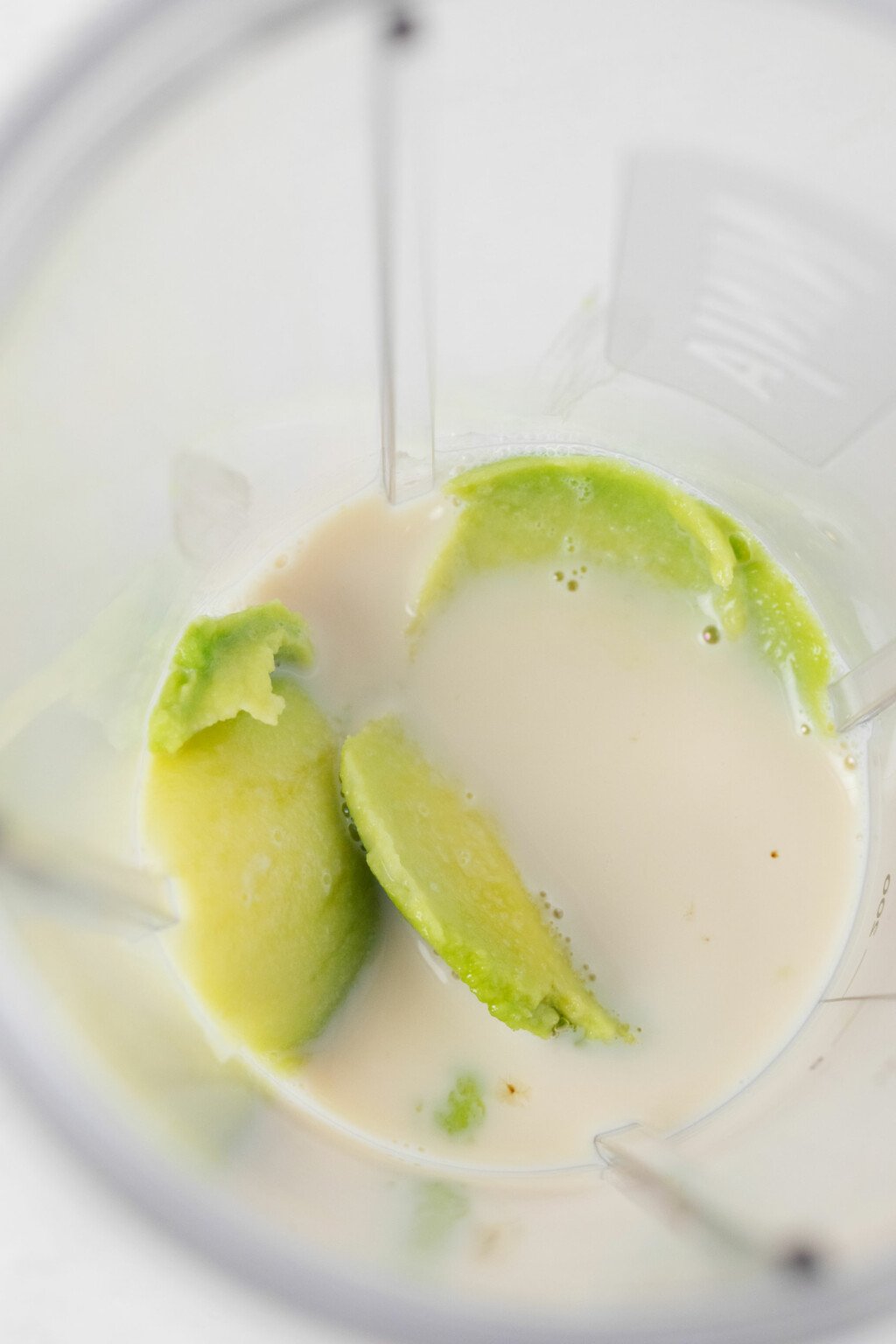 An overhead image of a avocado and non-dairy milk in the container of a mini blender.