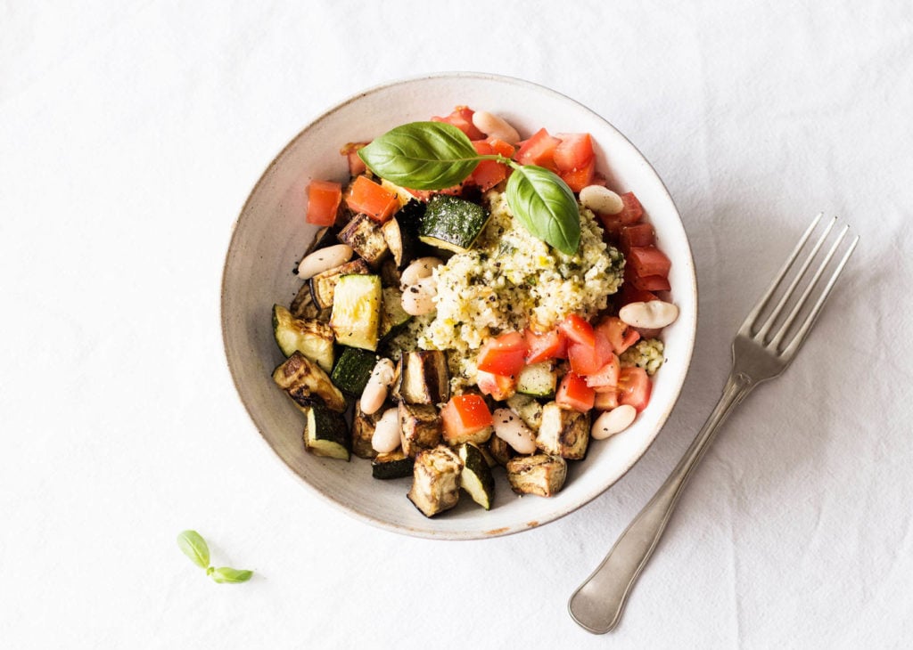 A white bowl has been filled with tomato zucchini eggplant whole grains and legumes It rests on a white cloth The Health Hop Vegan Meal Prep Dinner Ideas