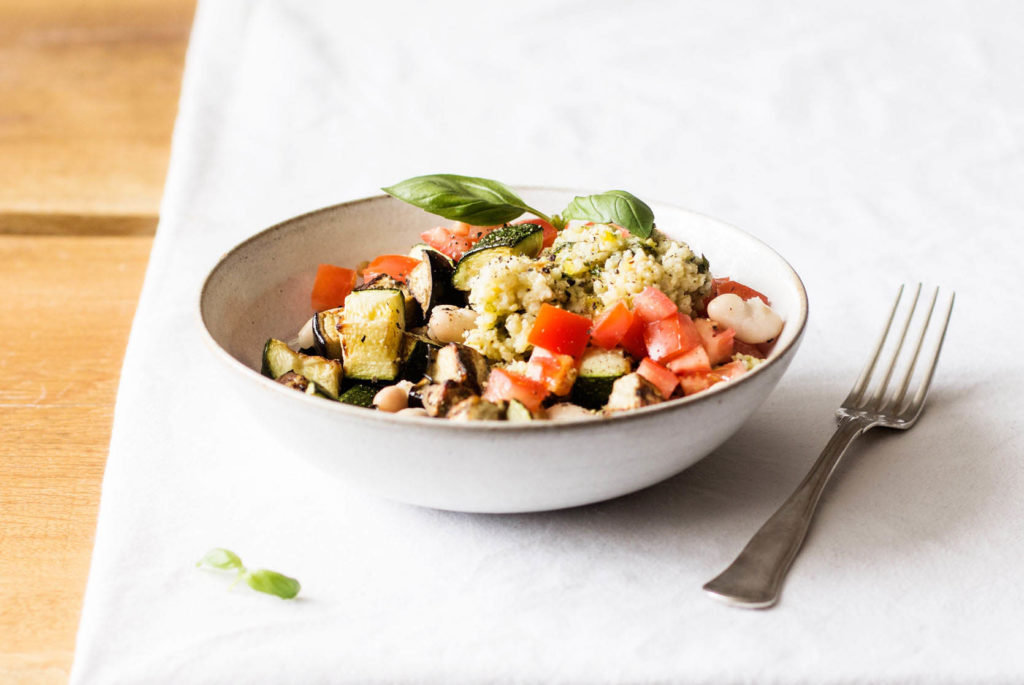 A pesto white bean bowl, made with summer tomatoes and eggplant, is resting on a white tablecloth with a fork nearby.