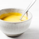 An angled photograph of a small, round ceramic bowl, which contains a golden colored sauce and a small serving spoon.