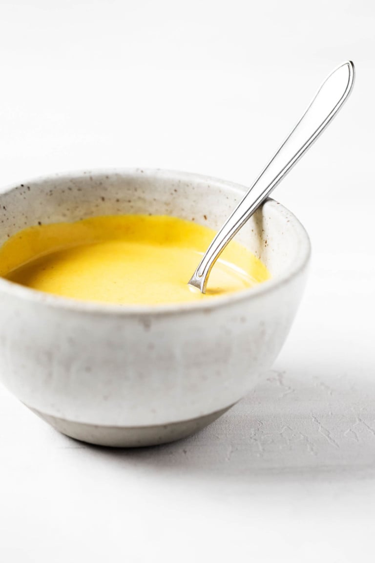 An angled photograph of a small, round ceramic bowl, which contains a golden colored sauce and a small serving spoon.