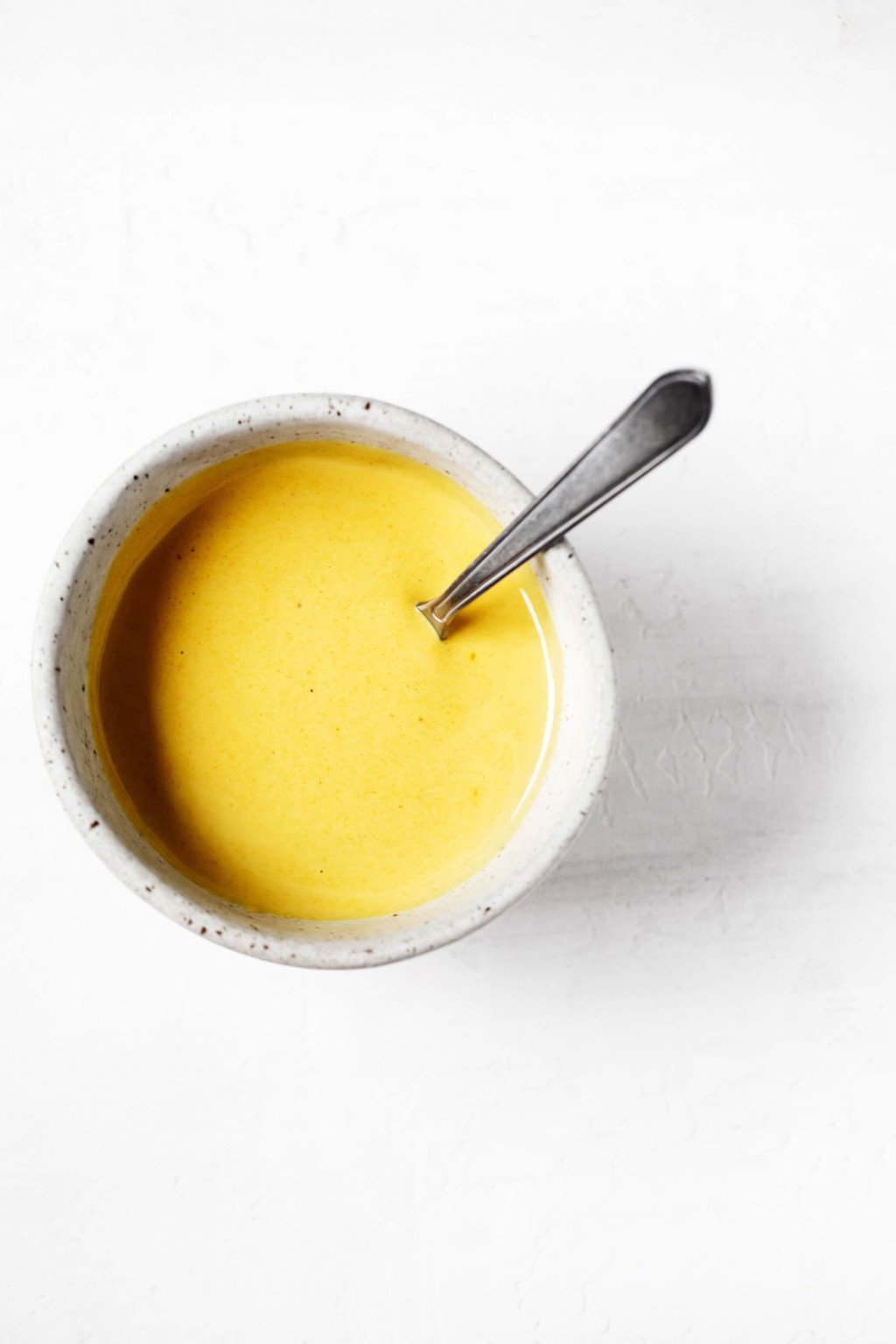 An overhead image of a small bowl containing a bright yellow colored dressing and a serving spoon.
