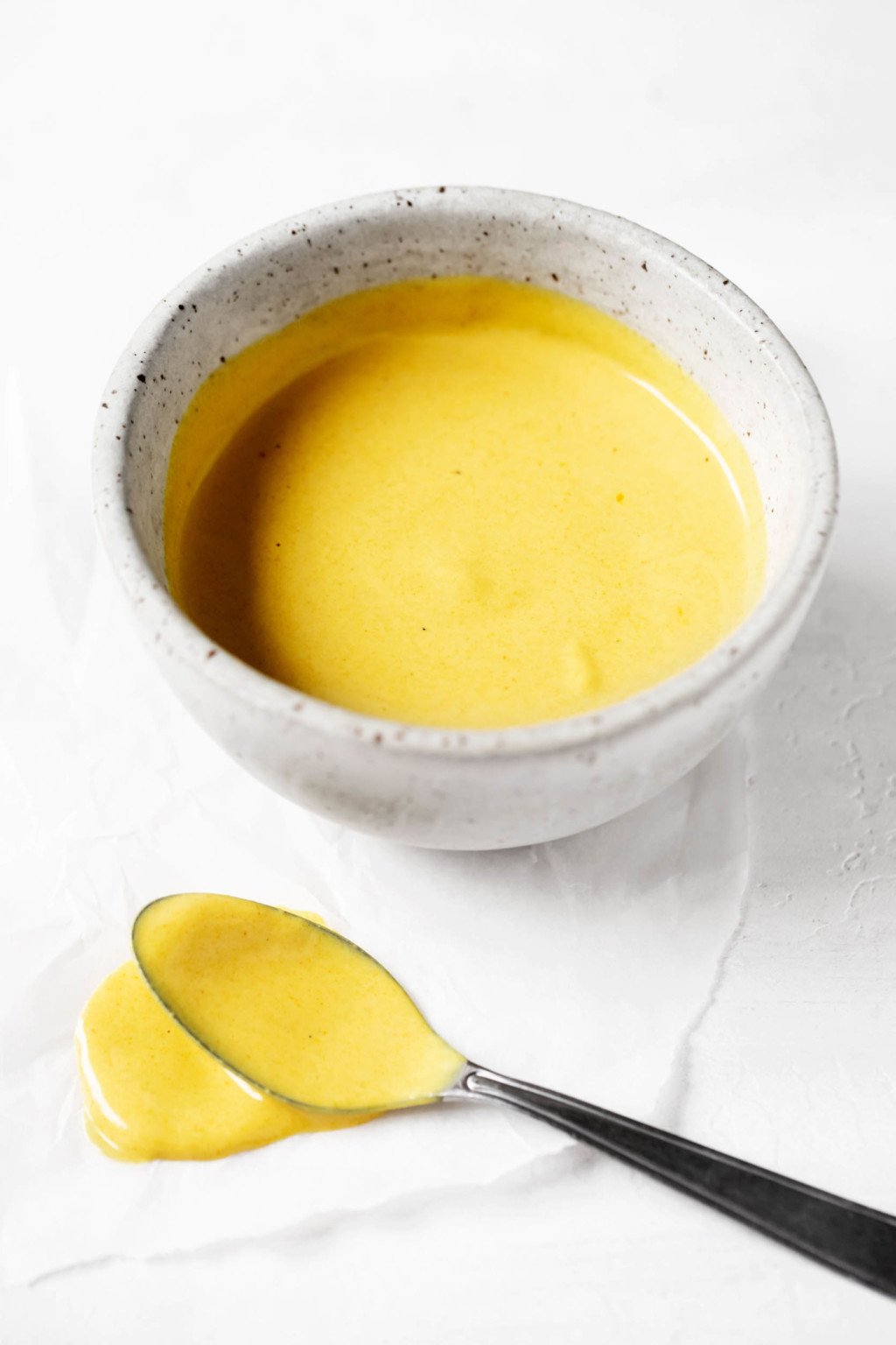 A small ceramic pinch bowl of turmeric tahini dressing has been laid out on a sheet of parchment. It's accompanied by a small serving spoon, which has recently been dipped in the dressing.