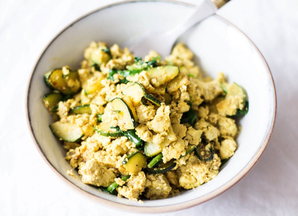 A round, white bowl has been filled with a mixture of scrambled tofu and thinly sliced, green zucchini.