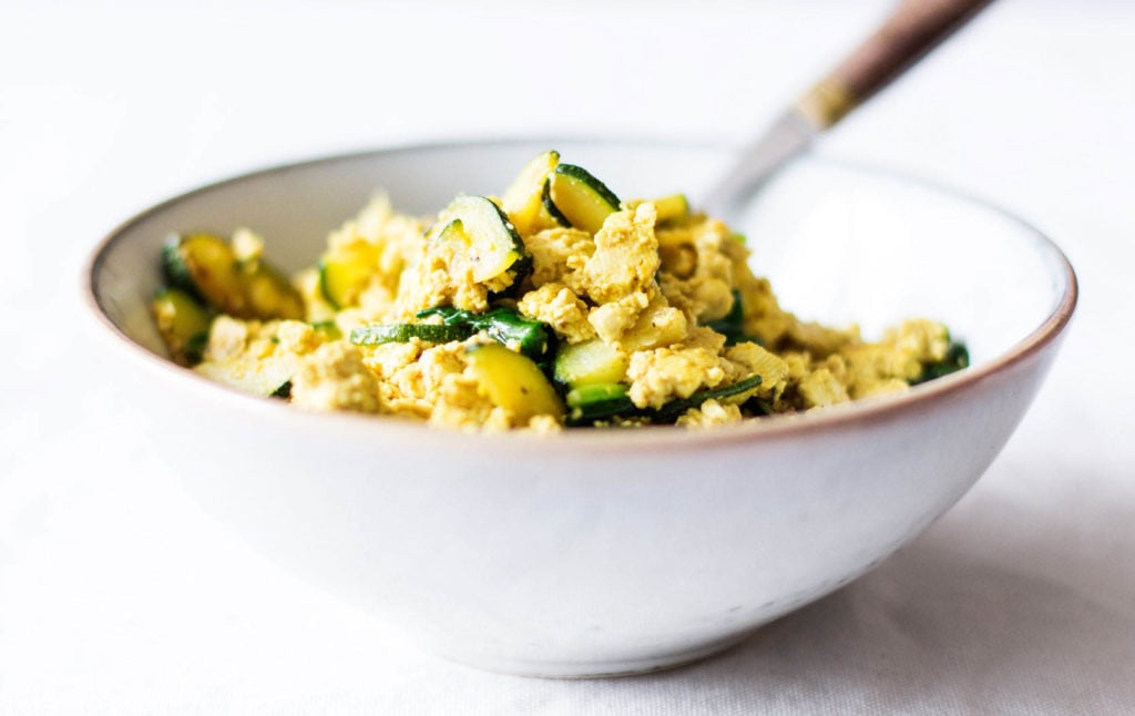 An angled photograph of a white ceramic bowl, which is filled with plant protein and zucchini. A serving fork pokes out of the food.