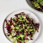 A white salad plate has been topped with a colorful, crunchy radicchio and Brussels sprout salad. It rests on a white surface.