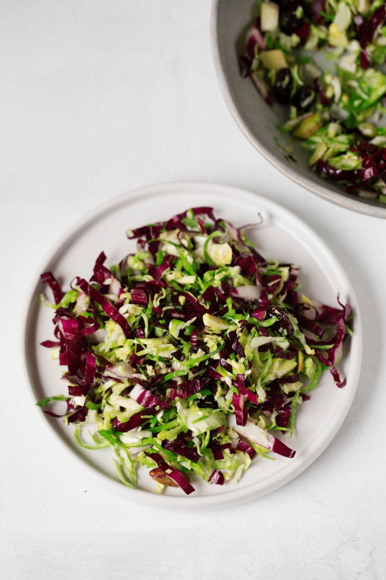 A white salad plate has been topped with a colorful, crunchy radicchio and Brussels sprout salad. It rests on a white surface.