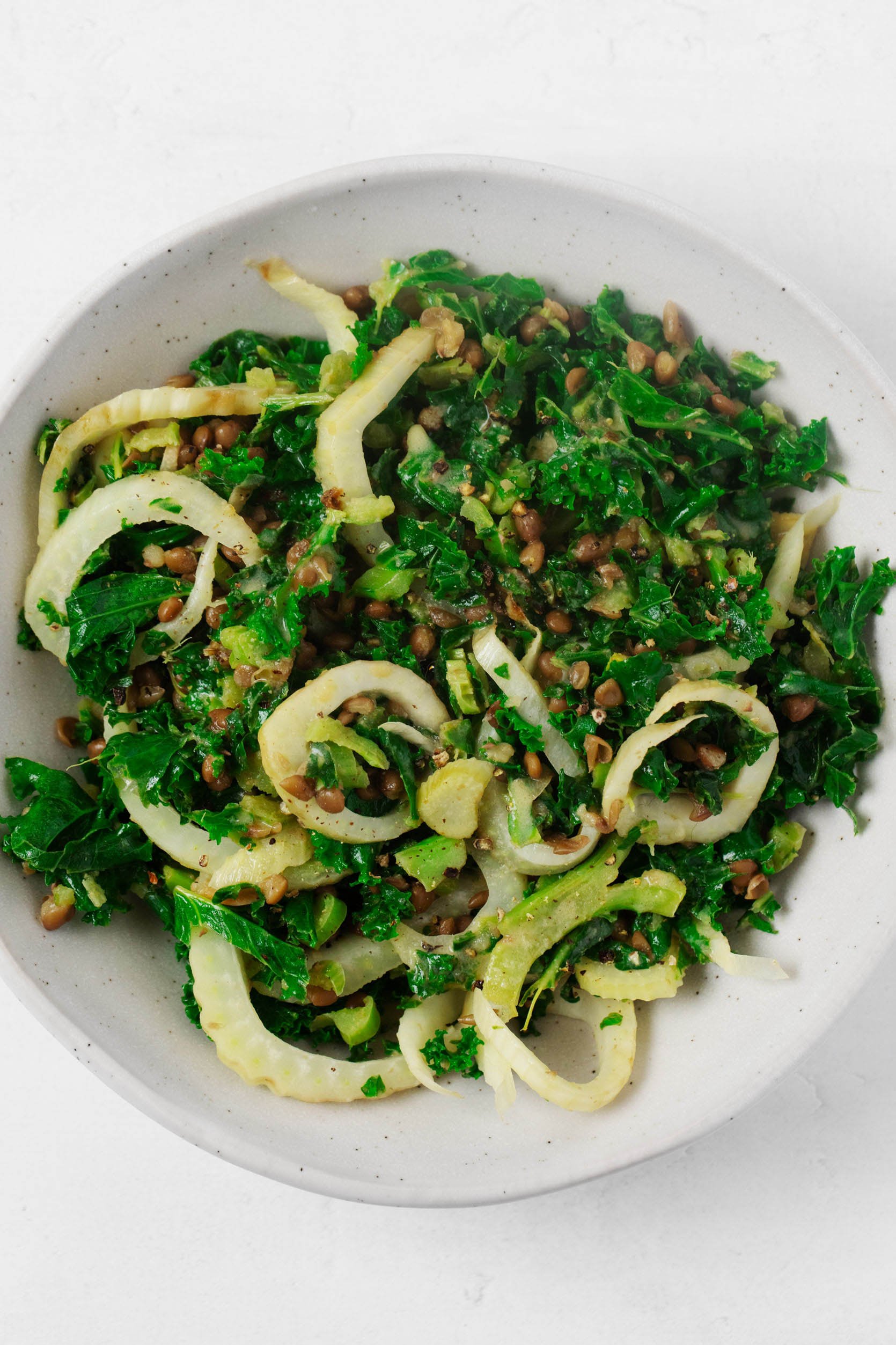 A dish of sautéed fennel has been mixed with warm kale, cooked lentils, and a vinaigrette. The dish is served in a white bowl.