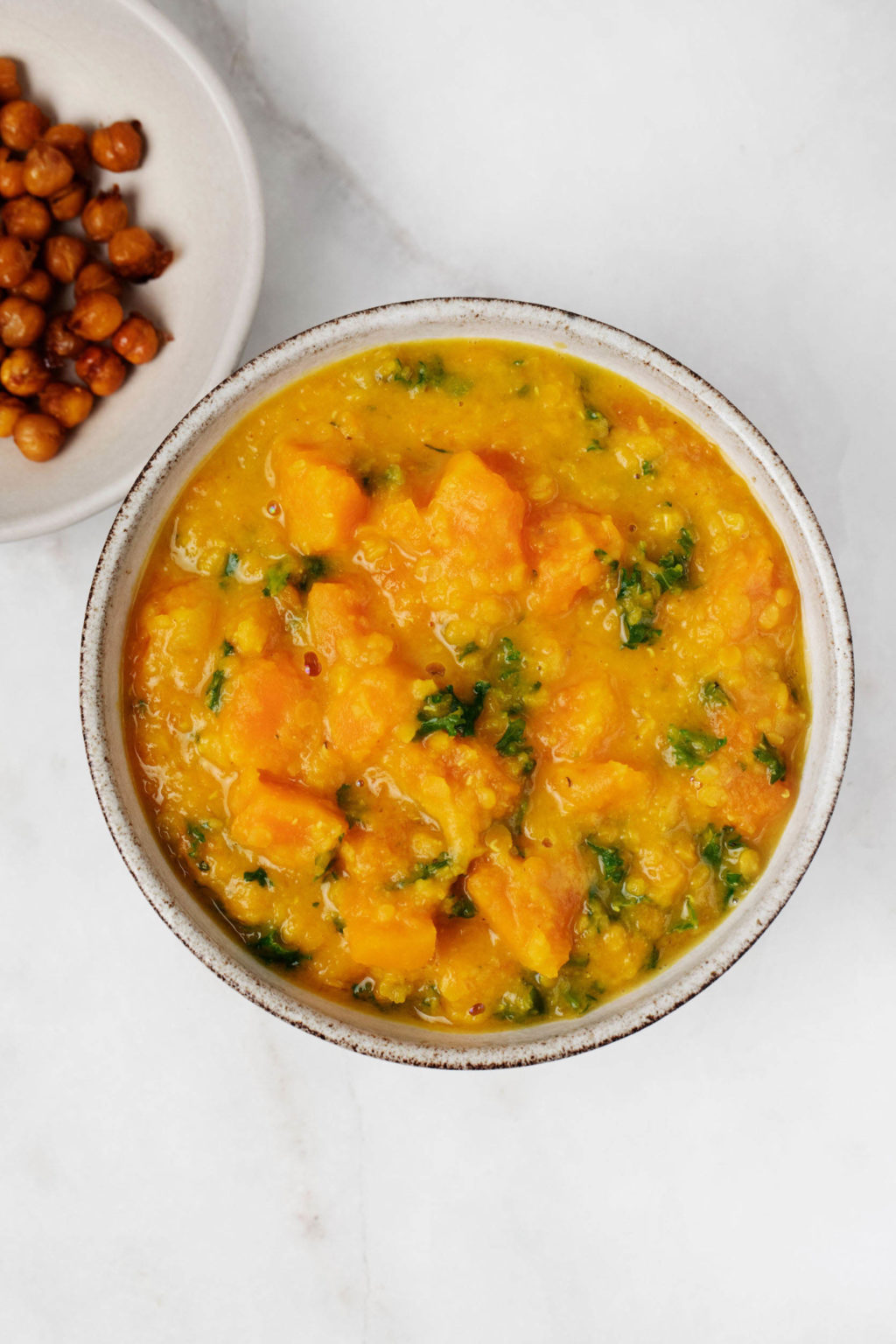 A bowl of nourishing vegetables and broth is standing next to a plate of toasted chickpeas on a white surface.