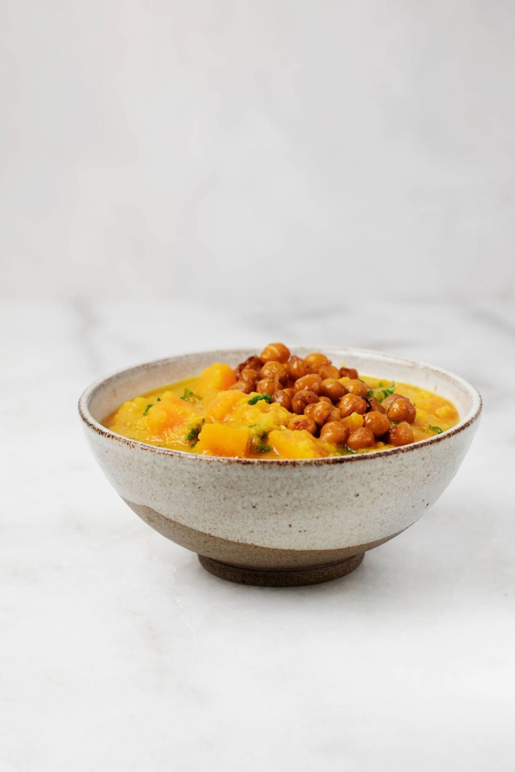 A white and brown ceramic bowl holds a portion of red lentil butternut soup. It rests on a white marble surface.