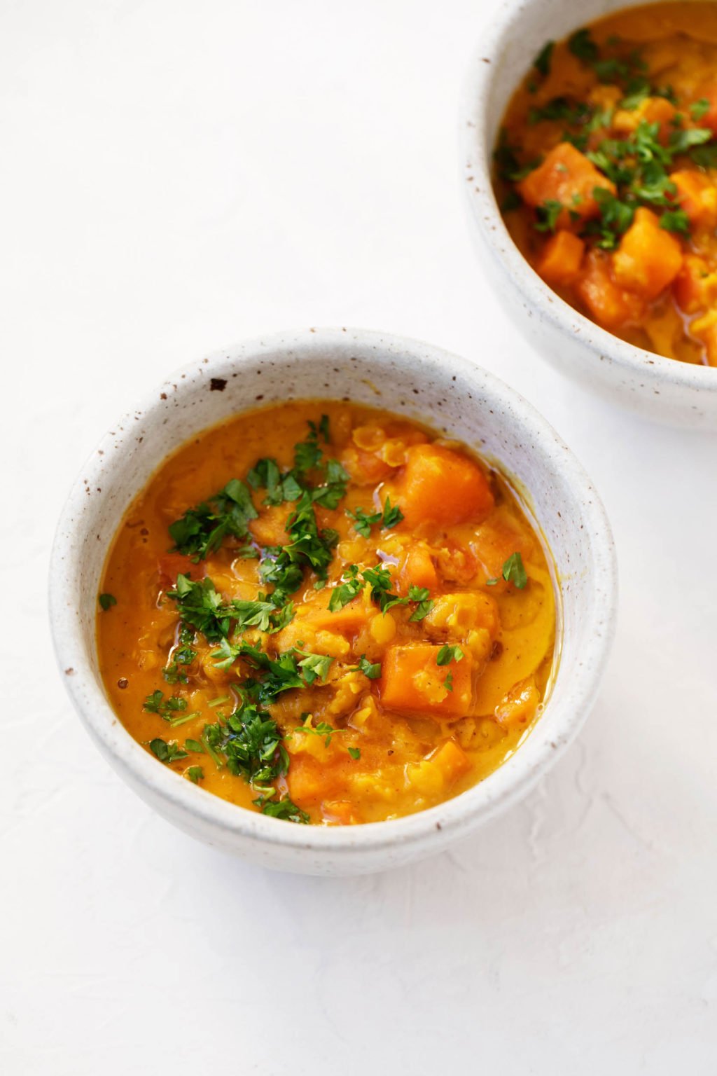 Two small, gray bowls have been filled with a plant-based, soupy dish of curried sweet potato lentils. Each bowl is garnished with chopped, fresh herbs.