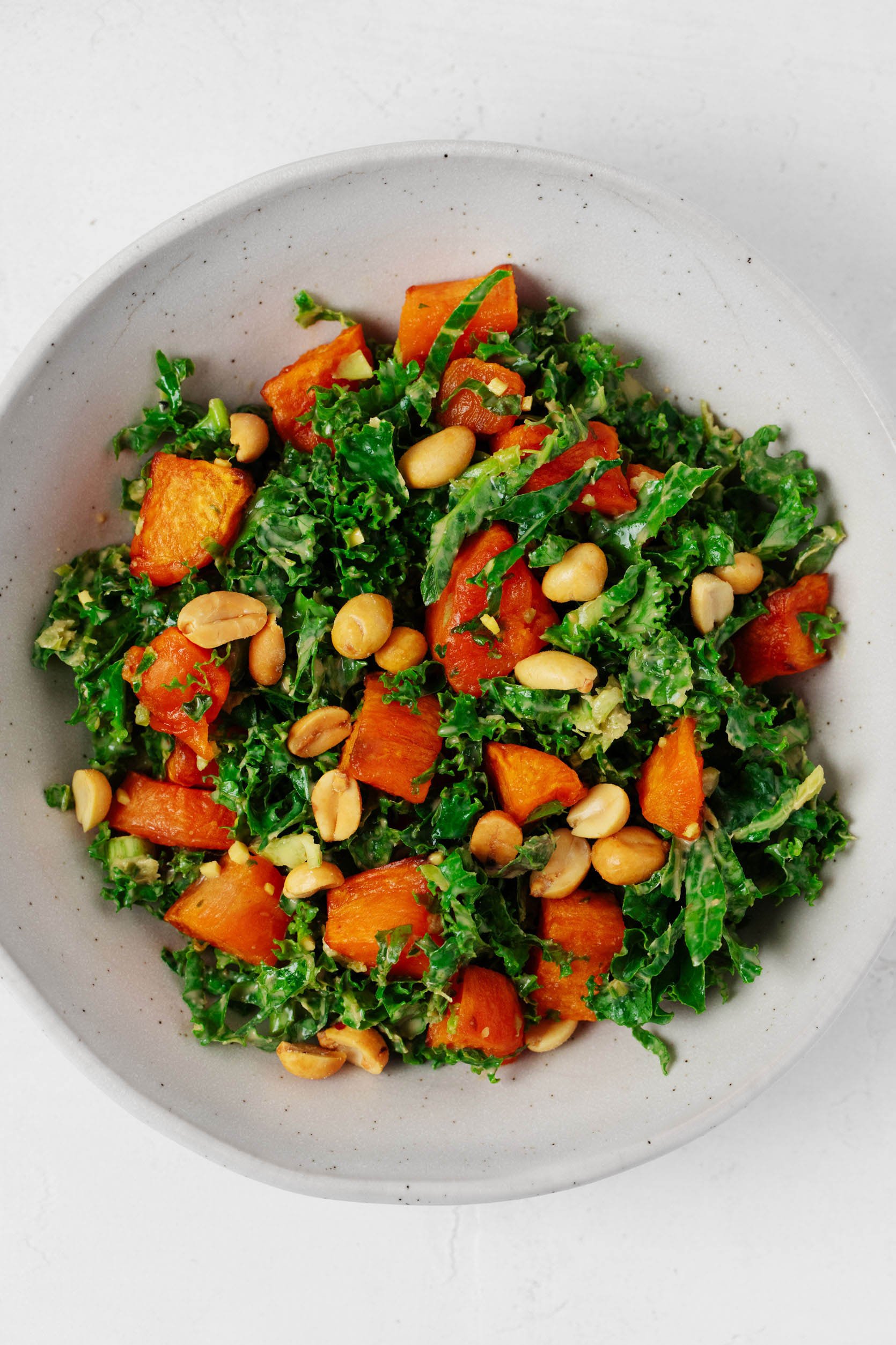 A spicy peanut kale salad with roasted sweet potato cubes is pictured in a large white mixing bowl.