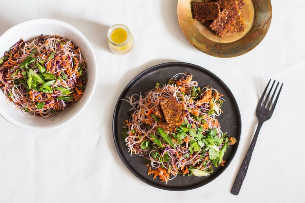 A vegan kelp noodle cabbage salad has been plated on a round, black matte plate. A small bowl of seared tempeh pieces rests to the top right. 