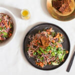 A vegan kelp noodle cabbage salad has been plated on a round, black matte plate. A small bowl of seared tempeh pieces rests to the top right.