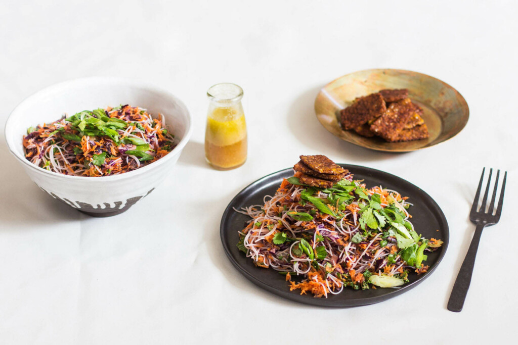 A colorful kelp noodle and tempeh salad has been plated and served with extra tempeh and a bright orange jar of salad dressing. Everything rests on a white tablecloth.