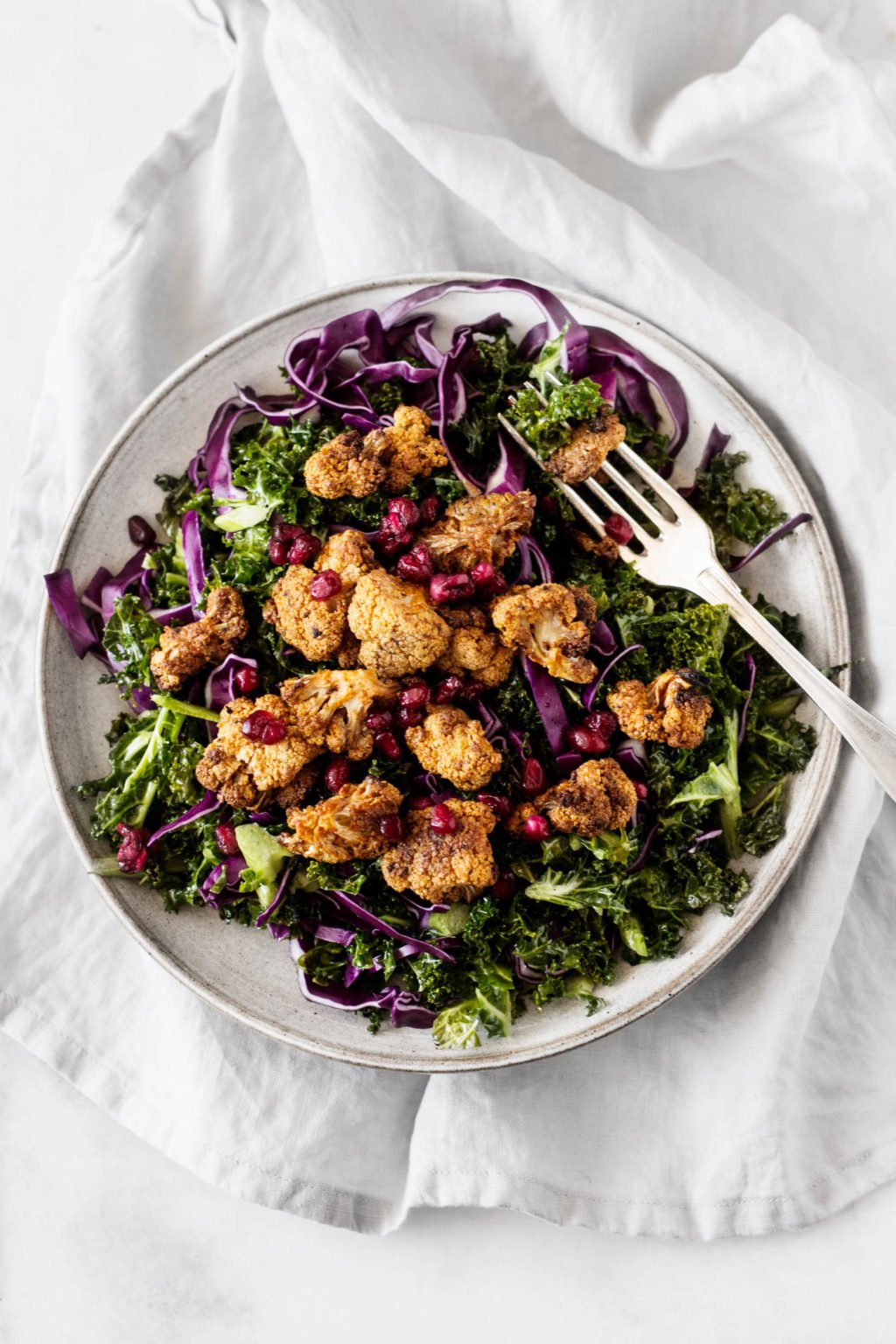 A serving plate is topped with vegan cauliflower pomegranate kale salad—a colorful mix of spice rubbed cauliflower, kale, purple cabbage, and pomegranate seeds.