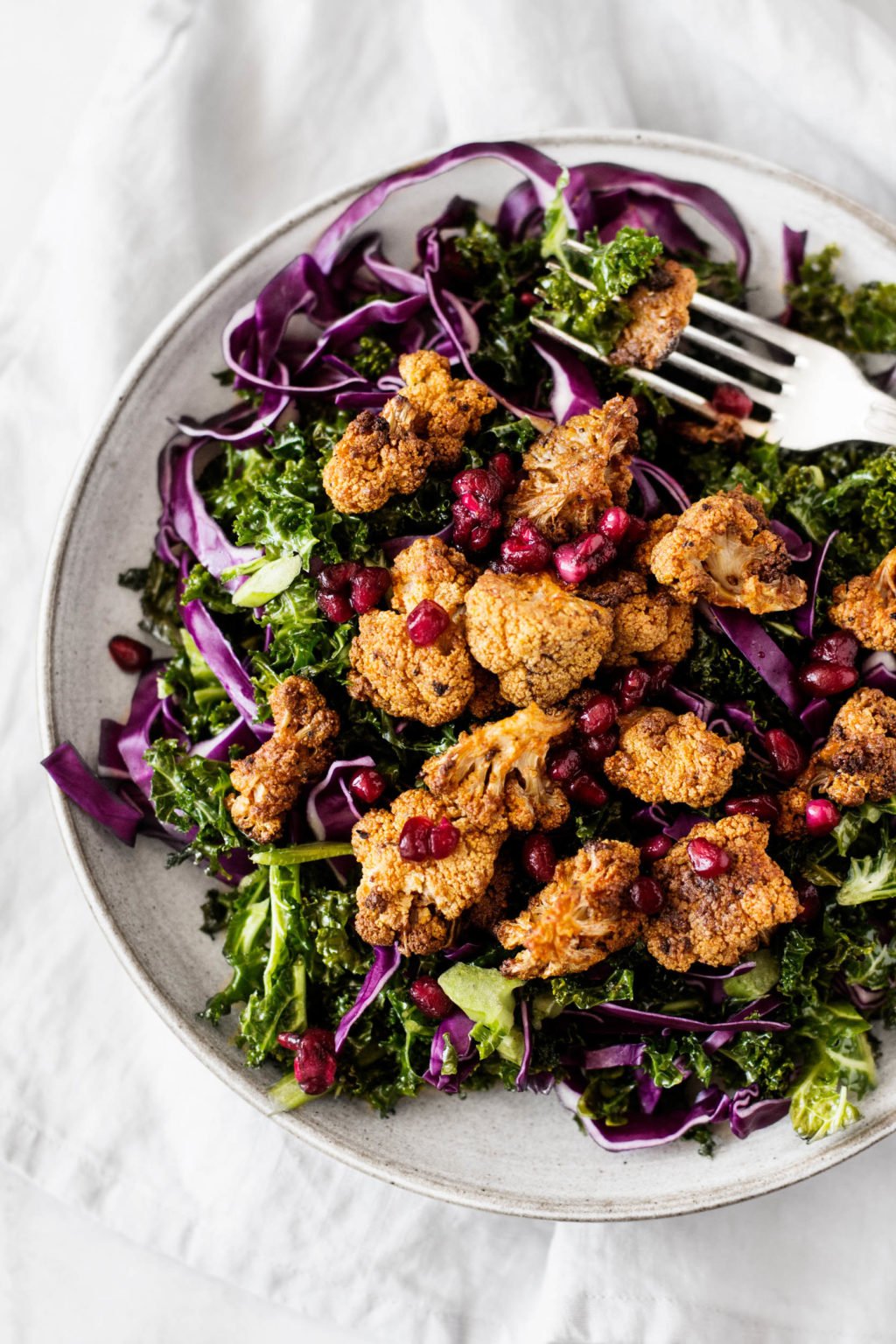 A shallow plate holds a festive, colorful salad of spiced, roasted cauliflower, raw kale, and pomegranate seeds.