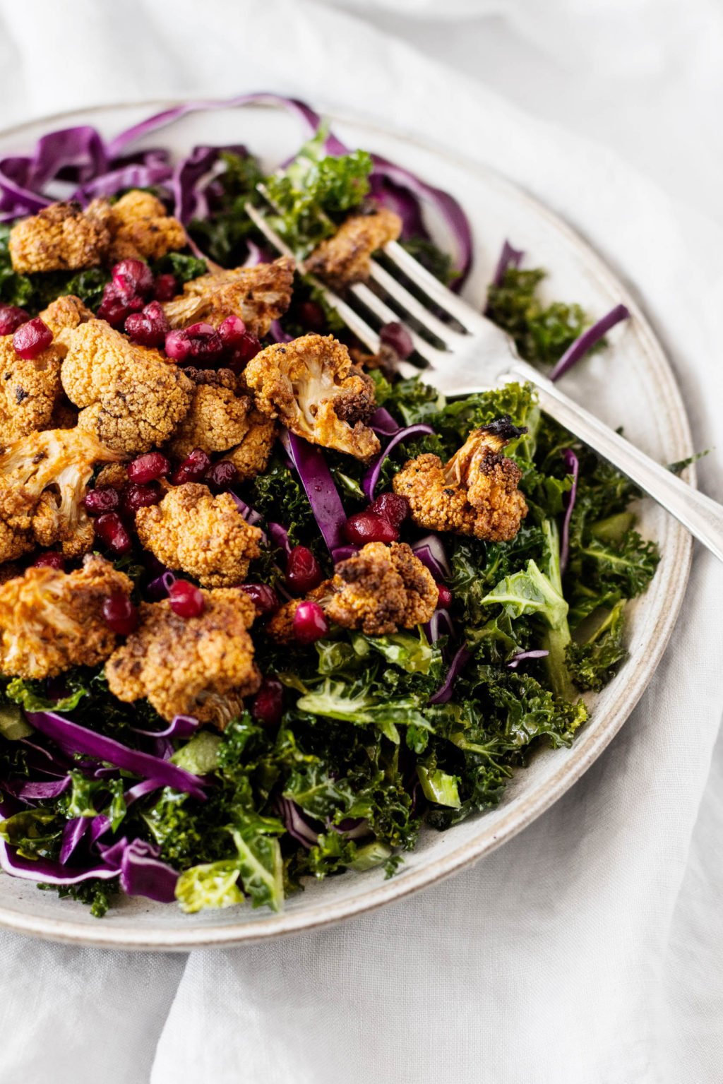Spice roasted cauliflower florets sit on top of a bed of kale, purple cabbage, and pomegranate seeds. All part of a festive vegan holiday salad.