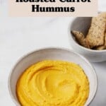 An overhead image of a white ceramic bowl holding an orange, creamy roasted carrot hummus. A small dish of crackers rests nearby.