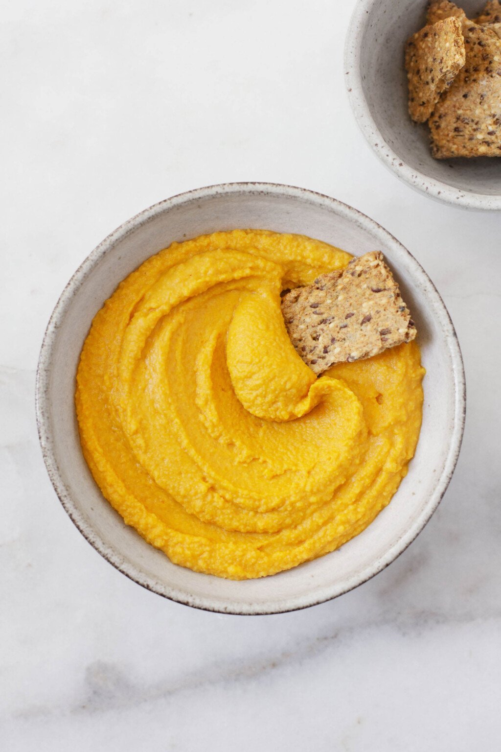 An overhead image of a white ceramic bowl holding an orange, creamy roasted carrot hummus. 