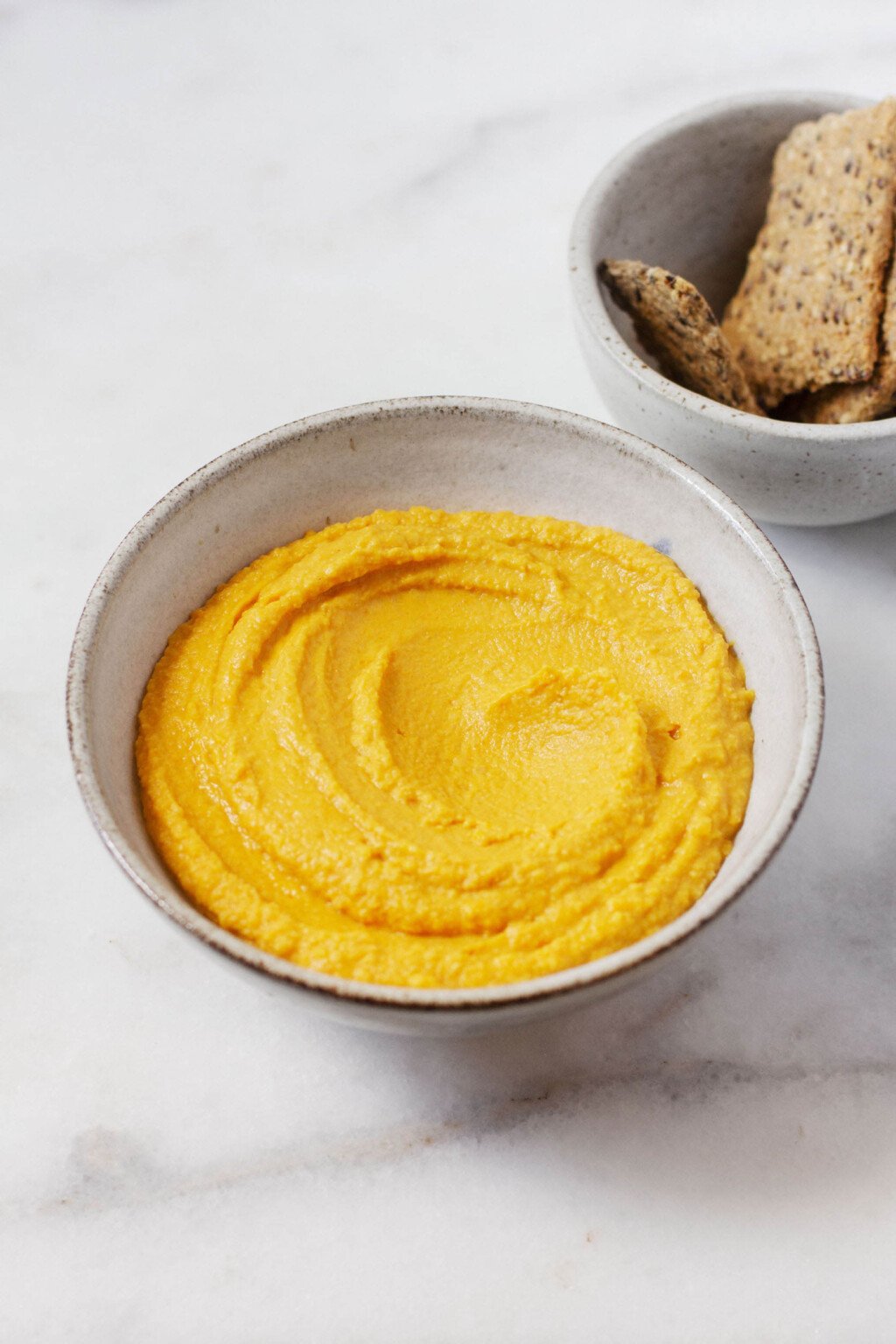 An overhead image of a white ceramic bowl holding an orange, creamy roasted carrot hummus. A small dish of crackers rests nearby.
