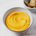 An overhead image of a white ceramic bowl holding an orange, creamy roasted carrot hummus. A small dish of crackers rests nearby.