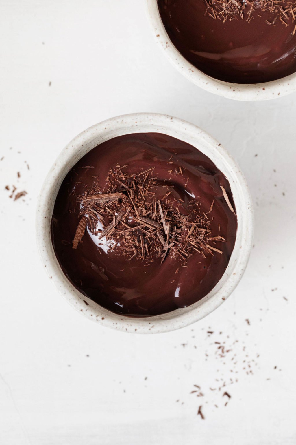 An overhead image of two small, ceramic bowls that are filled with a luscious, chocolate mixture. They rest on a white surface.